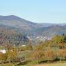 Blick über einen nach links abfallenden Hang mit Obstbäumen auf im Hintergrund aufsteigende, bewaldete Berge. Im Tal vor den Bäumen ist eine Siedlung zu erkennen.