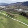 Blick aus der Luft auf welliges grünes Hügelland, durchzogen von Wegen und Hochspannungsleitungen. Rechts und im Hintergrund erheben sich bewaldete Berge. Davor breiten sich Siedlungen aus.