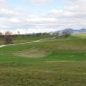 Blick auf nach innen gewölbte Wiesenhänge mit Wegen und kahlen Stellen im Mittelgrund. Links am Bildrand sind trichterförmige, von Bäumen umstandene Vertiefungen. Rechts im Hintergrund liegt eine Bergkette.