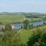 Von erhöhtem Standort aus blickt man über Baumwipfel auf eine flachhügelige Landschaft mit Wiesen, Äckern, Wald- und Rebflächen. Ein Fluss, links unterbrochen von einer Doppelschleuse, durchschneidet diese Landschaft von links nach rechts. 