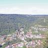 Blick aus großer Höhe auf bewaldete Berge sowie eine Siedlung, die im Vordergrund, am Fuß der Berge, liegt. Eine Bahnlinie durchschneidet den Ort. Rechts oben führt eine steile Bergstraße zu einer höher gelegenen Ortschaft.