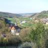 Von erhöhtem Standort aus blickt man auf ein schmales, an den seitlichen Rändern hügeliges und bewaldetes Tal. Eine kleine Ortschaft mit Kirche verteilt sich links und rechts darin.