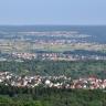 Von erhöhtem Standort blickt man auf eine von Wald umschlossene Ortschaft. Dahinter folgt eine Ebene mit Wiesen und Feldern sowie weiteren Siedlungen und Waldflächen.