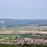 Von erhöhtem Standort schaut man auf eine weite Ebene mit Wiesen und Feldern, die sich am Fuße bewaldeter Berge ausbreiten. Im Vordergrund liegt eine Ortschaft an Waldrändern, links hinten ist eine größere Stadt erkennbar.