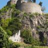 Aufwärts gerichteter Blick auf einen Teil der Festung Hohentwiel. Zu sehen ist ein vorgelagerter, oben eingebrochener runder Turm, der auf grauem, teils stark bewachsenem Vulkangestein sitzt.