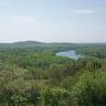 Blick von erhöhtem Standort auf eine weite Waldlandschaft mit kleineren Bergen im Hintergrund. Rechts vorne ist ein Felsplateau zu sehen, dahinter verläuft im Bogen ein breiter Fluss. Links unten sind Rebterrassen zu erkennen.