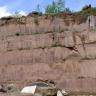 Blick auf eine hohe, rötlich graue Steinbruchwand. Das Gestein ist in mehreren Lagen blockhaft aufgeschichtet, mit fast glatter, leicht streifiger Oberfläche. Unten links liegen lose Blöcke. Die Kuppe oben ist bewachsen.
