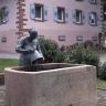Das Bild zeigt einen rechteckigen, aus rötlich grauem Stein gefertigten Brunnen. In den Stein sind Zeichen und Buchstaben eingeritzt. Eine Frauenfigur mit Wassereimer ergänzt den Brunnen. Im Hintergrund steht ein Gebäude, ebenfalls aus rötlichem Stein.