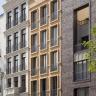 Das Bild zeigt die dreifach unterteilte Fassade eines mehrstöckigen Wohnhauses mit zahlreichen Fenstern. Die Fassaden bestehen aus grauem Klinker rechts sowie hellbraunen und weißen Steinplatten (mitte und links).
