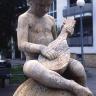 Die Aufnahme zeigt eine Steinfigur, sitzend auf einer Steinkugel. Figur und Kugel sind aus gelblichem bis grauem Material; die Figur ist einem Jungen nachgebildet, der ein Zupfinstrument in den Händen hält.