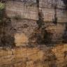 Blick auf eine hohe, unten rötlich braune, oben graue Steinbruchwand. Die Wand ist von Spalten und höhlenartigen Vertiefungen durchzogen.