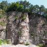 Blick auf eine hohe, rötlich graue Steinbruchwand. Die Wand ist mehrfach eingekerbt, die Kuppe oben ist bewaldet. Zwischen dem Gestein wachsen auch Büsche.