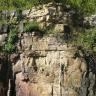 Blick auf eine hohe Steinbruchwand aus rötlich braunem bis grauem Gestein. Im oberen Teil wachsen Sträucher, auf der Kuppe wachsen Bäume.