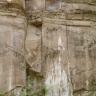 Blick auf eine hohe Steinbruchwand. Im oberen Teil befinden sich dünnplattige Abraumschichten, getrennt durch größere Blöcke. Darunter steht eine von senkrechten Rissen durchzogene Kluftwand an.