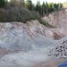 Steinbruchwand mit ocker- bis rotgrauem, absandendem Granit im linken Abschnitt und kompaktem rotgrauem Granit im rechten Abschnitt. Oberhalb der Steinbruchwand liegt ein Mischwald. Im Vordergrund rechts liegt ein Haufen mit Steinen aus dem Granitbruch.