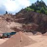 Abbauwand eines Steinbruchs mit einheitlich rotgrauem Granit. Im Vordergrund ein mobiler Brecher und mehrere Haufwerke. Rechts, oberhalb des Steinbruchs, liegt ein Mischwald.