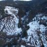 Blick aus der Vogelperspektive auf einen teilweise schneebedeckten Hang, der vor allem am oberen Ende bewaldet ist. Im Zentrum des Bildes ist ein Bach zu erkennen, der stark angeschwollen den Hang hinabfließt.