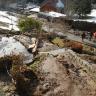 Blick von oben herab auf ein Grundstück, das von Schlamm, kleinen Steinen und Holzresten bedeckt ist. Dahinter ist eine Straße zu sehen, neben der größere Gesteinsbrocken liegen. Im Hintergrund stehen weitere Häuser.