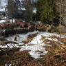 Das Bild zeigt einen stark angeschwollenen Bach, der an einem Gebäude entlang auf eine Brücke zufließt. Die Brücke ist mit Schlamm, Steinen und Ästen bedeckt. Dahinter stehen zwei Bagger und einige Personen auf einer Straße; im Hintergrund weitere Häuser.