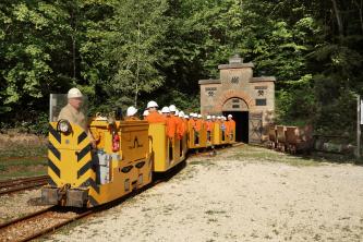 Blick auf die Grubenbahn eines ehemaligen Bergwerkes. Die gelben Wagen sind mit Besuchern besetzt, die orange Jacken und weiße Helme tragen. Im Hintergrund der mit Zinnen bekrönte Eingang des Bergwerkes.