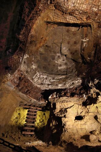 Blick in das Innere eines Besucherbergwerkes. Gezeigt wird ein hoher Raum mit hellem Gestein unten und rötlich braunem Gestein oben. Oben sind teilweise Eisengitter angebracht. Unten links ist eine kleine Leiter aus Eisenschienen zu erkennen.