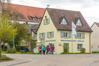 Blick auf ein älteres, kleines gelbes Haus mit blaugrauen Fensterläden. Es hat einen überdachten Eingang und trägt eine Inschrift: Naturschutzzentrum Federsee. Vor dem Eingang steht eine Besuchergruppe.