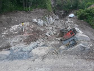 Das Bild zeigt ein Bachbett, in dem gerade kein Wasser fließt. Große Steinblöcke säumen das Ufer, im Bachbett befindet sich Geröll und kleinere Steinblöcke. Am unteren Bildrand verläuft ein neu befestigter Weg. Am Rand liegt eine Baggerschaufel.