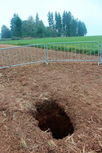 Im Vordergrund dieses Bildes ist ein auf einem Acker entstandenes Erdloch zu sehen. Das Erdloch ist mit Gittern abgesperrt. Im Hintergrund ist ein Waldstück zu erkennen.