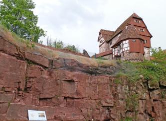 Blick auf eine rötlich graue, ehemalige Steinbruchwand. Der obere Rand des dickbankigen Gesteins ist mit Netzen gesichert. Rechts ist ein verwinkeltes Fachwerkgebäude mit vorgelagertem Turm zu sehen, darunter ist das Gestein stark zugewachsen.