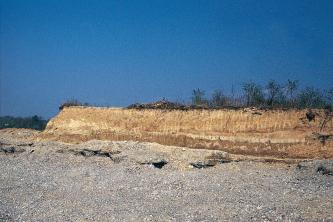 Das Bild zeigt eine graue Schotterfläche, über der sich wie eine Insel rötlich braunes Gestein erhebt. Die Kuppe der Insel ist rechts dünn bewachsen.