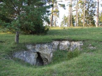 Das Bild zeigt eine ovale Vertiefung inmitten einer Waldlichtung. Neben gelblich grauen Felsen ist am unteren Rand auch eine kleine Höhle erkennbar.