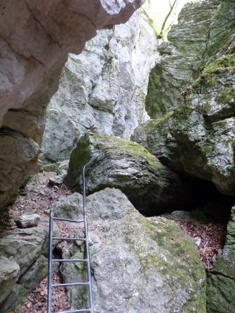 Das Bild zeigt eine enge Felsenschlucht mit begehbarem Pfad. Das Gestein der Felswände rechts und im Hintergrund ist grünlich grau. Einige größere Felsbrocken liegen im Schluchteingang. Links ist eine Aufstiegsleiter sichtbar.