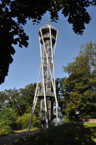 Das Bild zeigt einen Aussichtsturm, der zwischen Bäumen aufragt. Der Turm, der in der Form einer Sanduhr ähnelt, hat Treppen, Geländer und Stützen aus Metall.