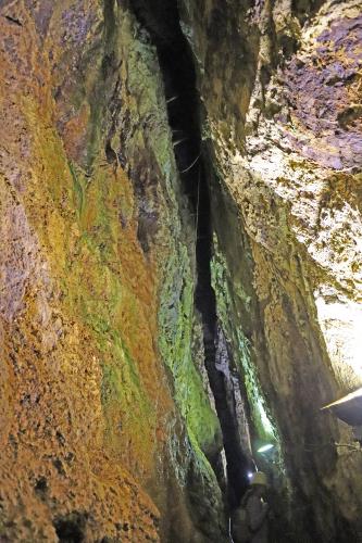 Das Bild zeigt eine senkrecht verlaufende Spalte zwischen Gesteinswänden. Das Gestein um die Spalte herum ist grünlich verfärbt. Links außen, zum Vordergrund hin, sind rostbraune Farben vorherrschend.