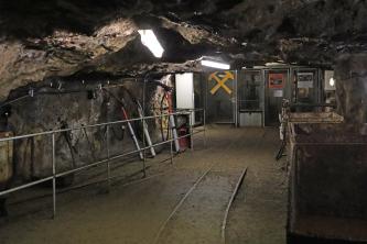 Blick in einen Stollen des Besucherbergwerks Teufelsgrund. Links sind Abbauwerkzeuge ausgestellt, rechts stehen alte Förderwagen an der Wand. Schienen auf dem Stollenboden führen zu einem vergitterten Blindschacht im Hintergrund.