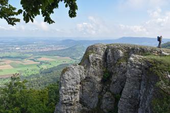 Blick von erhöhtem Standort auf eine rechts vorspringende Felsengruppe, die als Aussichtspunkt dient. Von hier schaut man auf Felder, bewaldete Hügel und Ortschaften herab. Im dunstigen Hintergrund sind noch einige Höhenzüge erkennbar.
