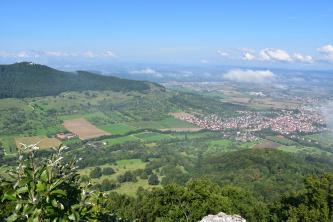 Blick aus großer Höhe über eine weite Landschaft mit Feldern und bewaldeten Hügeln. Rechts verteilen sich mehrere Ortschaften. Nach links hin erhebt sich ein bewaldeter Bergrücken mit einer Burg.