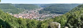 Blick von erhöhtem Standort auf ein stark besiedeltes Tal. Links und rechts der Bebauung erheben sich bewaldete Berge mit flachen Kuppen. Auch im Vordergrund steigen zu den Seiten hin bewaldete Berge auf. Rechts sind zudem Felsvorsprünge zu sehen.