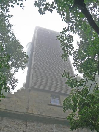 Zwischen belaubten Bäumen ragt ein hoher Turm in einen nebligen Himmel. Der Turm hat eine Basis aus Mauer- und Backsteinen sowie ein Fenster. Der eigentliche Turm besteht aus sowohl senkrecht wie auch waagrecht verbauten Holzbalken.