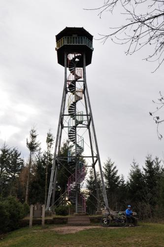 Blick auf einen überdachten, hohen Aussichtsturm aus Metall. Der Turm ist offen und hat eine innen hinaufführende Wendeltreppe, ebenfalls aus Metall. Der Turm steht auf einer Anhöhe, hinter ihm sind Bäume, über die er aber weit hinausragt.