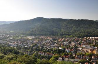 Blick von sehr hohem Standort auf die Häuserzeilen einer größeren Stadt, die sich bis zu einem bewaldeten Bergrücken im Hintergrund ausdehnen. 