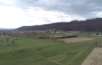 Blick von oben auf flache Wiesen und Äcker, durchkreuzt von Wegen, einem von Bäumen gesäumten Graben sowie einer Straße. Links und mittig folgt eine Siedlung, nach rechts erhebt sich ein bewaldeter, langgezogener Bergrücken.