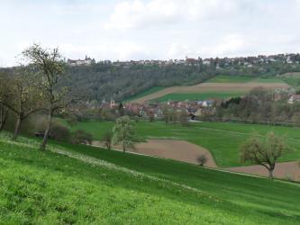Der Blick geht über eine nach unten hin abfallende grüne Wiese über ein bewirtschaftetes Tal, an das sich ein wieder ansteigender, teils bewaldeter Hang anschließt. Im Mittelgrund sowie auf der Anhöhe liegen Ortschaften, hinten links auch ein Schloss..
