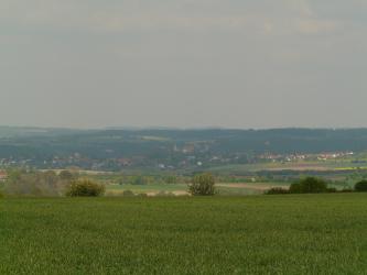 Hinter einer Grünlandfläche sieht man eine weite Hochebene, die zur Mitte hin leicht vertieft ist. Äcker und Wiesen wechseln sich ab mit Ortschaften und größerer Bewaldung im Hintergrund.