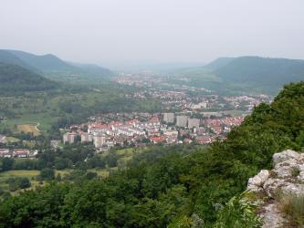 Von erhöhtem Standpunkt aus hat man - über Felsen und Baumwipfel - einen weiten Blick auf ein Tal und mehrere, fast zusammenhängende Ortschaften, die bis zum Horizont reichen. Links und rechts grenzen bewaldete Höhenzüge an.