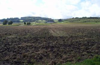 Blick über weite, dunkelbraune Äcker, die bis zum Hintergrund reichen. Dort schließt sich eine tiefer liegende, begrünte Landschaft an mit bewaldetem Hügel links und einem zweiten, von Gebüsch durchsetzten Hügel rechts.