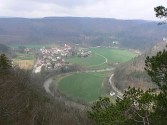 Von erhöhtem Standpunkt aus blickt man auf eine flache, teils besiedelte und teils grüne Talaue, die ein Fluss C-förmig durchquert. Steile, bewaldete Höhenzüge umrahmen die Aue. Im Zentrum der Siedlung liegt ein Kloster.