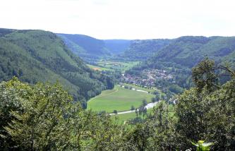 Von hohen bewaldeten Steilhängen umschlossen, liegt eine breite grüne Ebene in der Bildmitte. Ein im Bogen verlaufender Fluss durchquert die Ebene, die sich zum Hintergrund hin fortsetzt. Eine Siedlung liegt am Fuß der Anhöhe rechts.