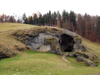 Auf einem grünen, nach rechts abfallenden Hang sitzt ein mächtiger, mit Gras und Moos bewachsener, grauer Felsen. Rechts weist der Fels eine Höhlenöffnung auf, die in die Tiefe führt. Im Hintergrund steht Wald.