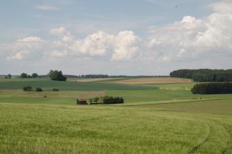 Blick über eine grüne Wiese auf wechselseitig ansteigendes, hügeliges Gelände mit Wiesen, Äckern und Waldflächen.